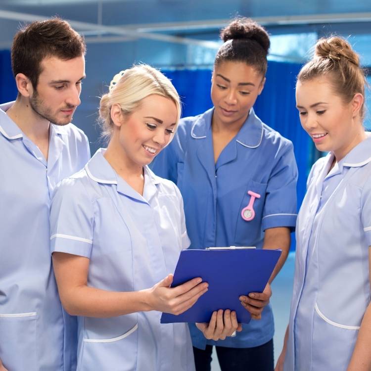 Nursing assistants in blue scrubs attentively discussing a patient's vitals on a hard board, exemplifying dedication and care.